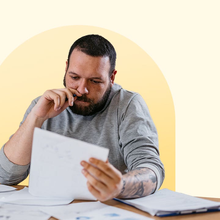 Man thoughtfully reviewing a document at a table that has many papers on it. The image has a light yellow background with a darker yellow circle behind the man.